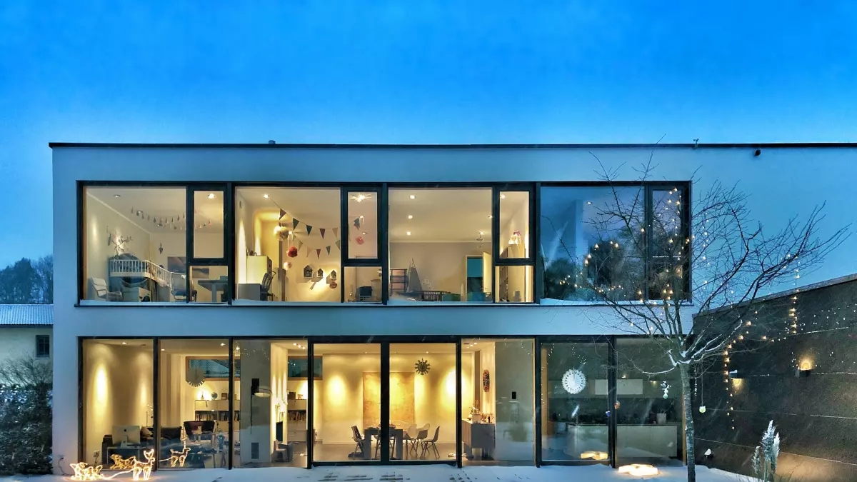 A modern house with large windows showcasing the interior, taken at dusk with the exterior lights on, showing a cozy interior and a snowy garden. 